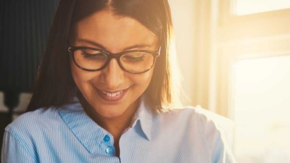 Young smiling woman in light blue shirt wearing glasses, sat in a sunlit lounge, looking at laptop