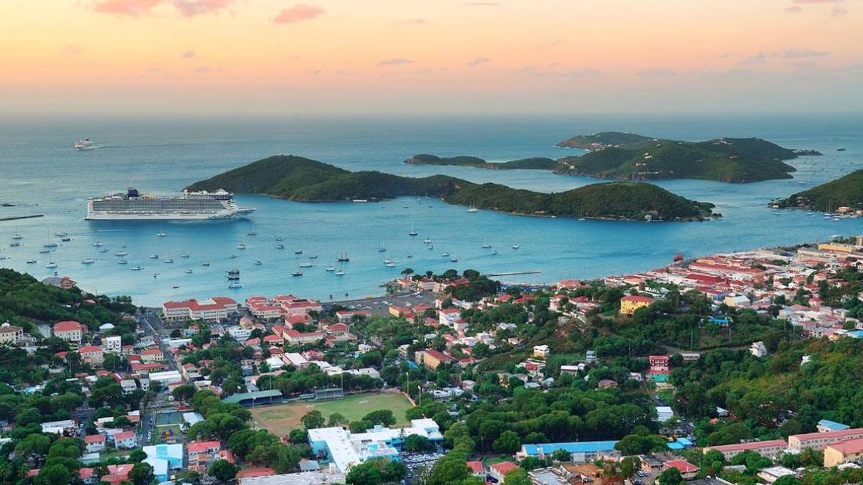 Aerial view of a coastal town with a large cruise ship docked near small islands, surrounded by buildings, roads, green spaces and boats, under an orange and pink sky 