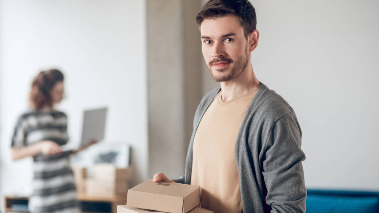 Man holding cardboard parcels and smiling at the camera, with a woman in the background looking at a laptop in a home setting.