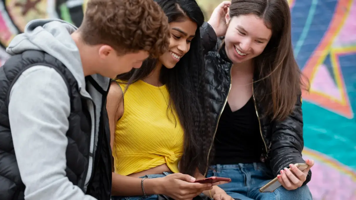 Three teens sit in front of a bright graffiti wall, checking something out on a phone together 