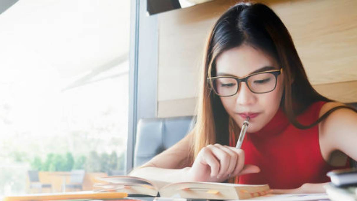 Woman sat in a well-lit café, holding pen to her mouth while reading from a book for her studies
