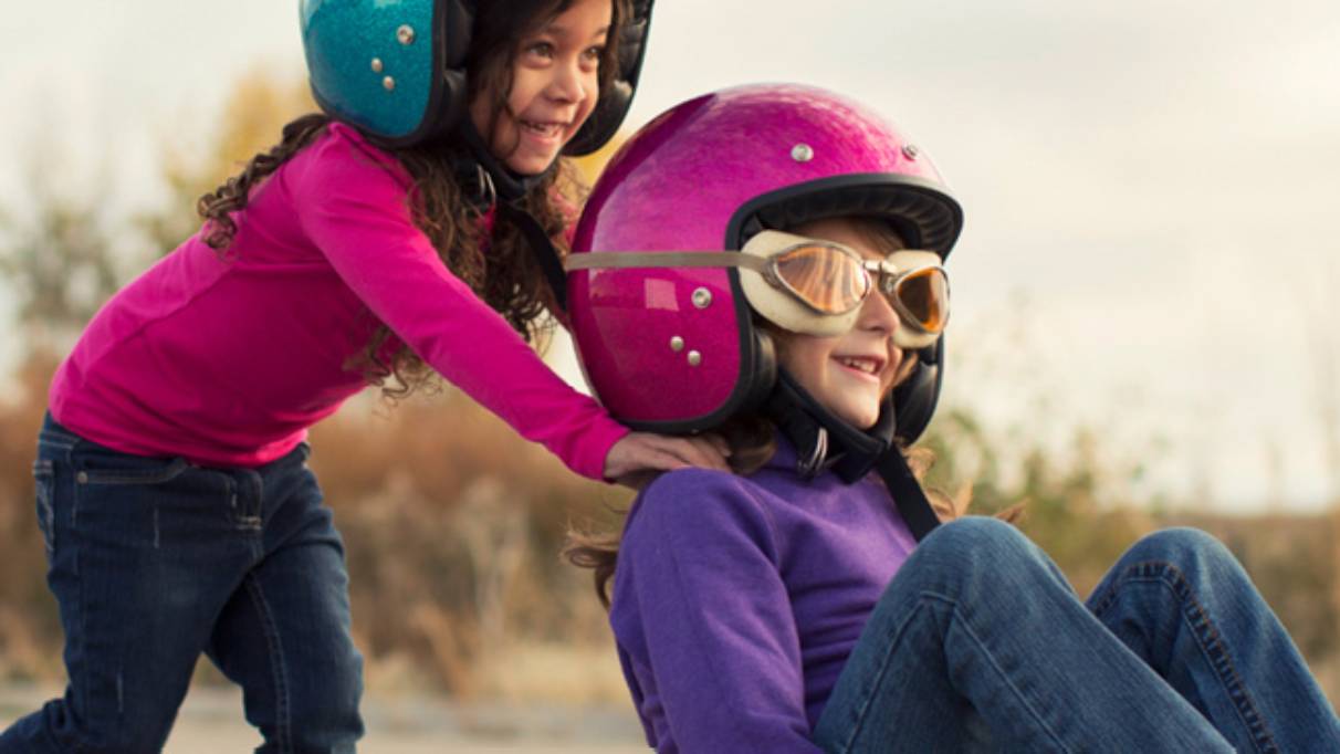Two smiling girls wearing helmets, one sat on skateboard wearing goggles, being pushed by the other