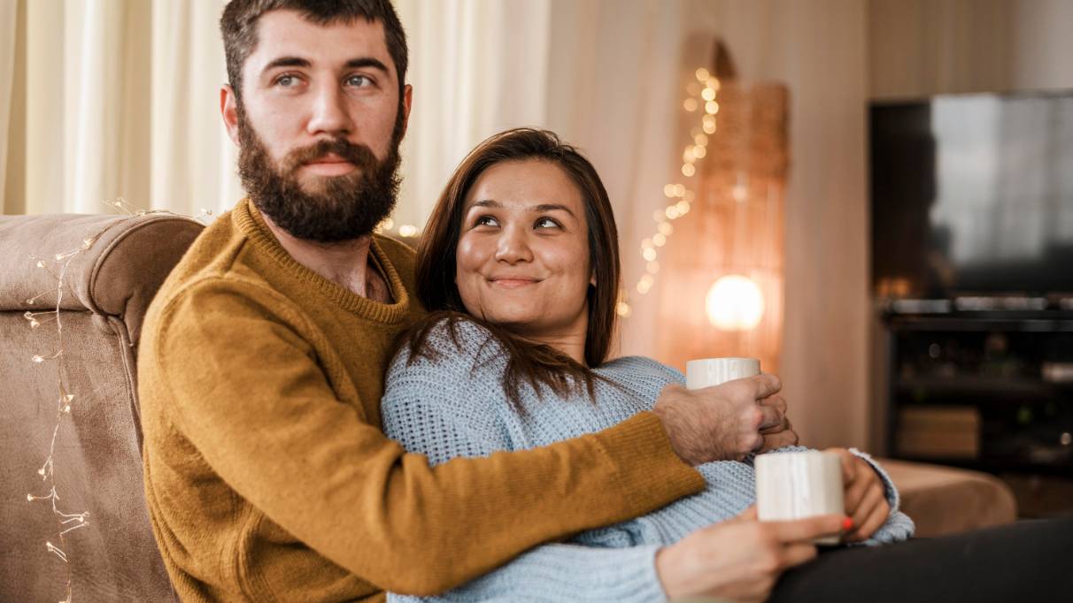 A couple sit at home, arms around each other and holding coffee cups