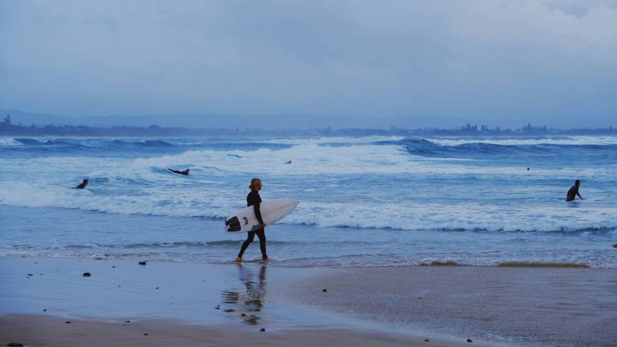 A surfer carries her board along the beach were the water laps the sand. In the background, three other surfers ride frothy waves