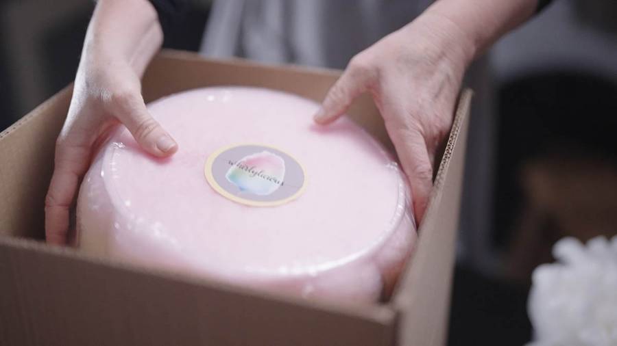 A pair of hands placing a round, sealed, pink candyfloss cake labelled 'Whirlylicious' into a cardboard box