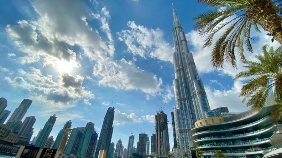 Dubai skyline with Burj Khalifa, modern buildings, outdoor seating, and a palm tree under a partly cloudy sky