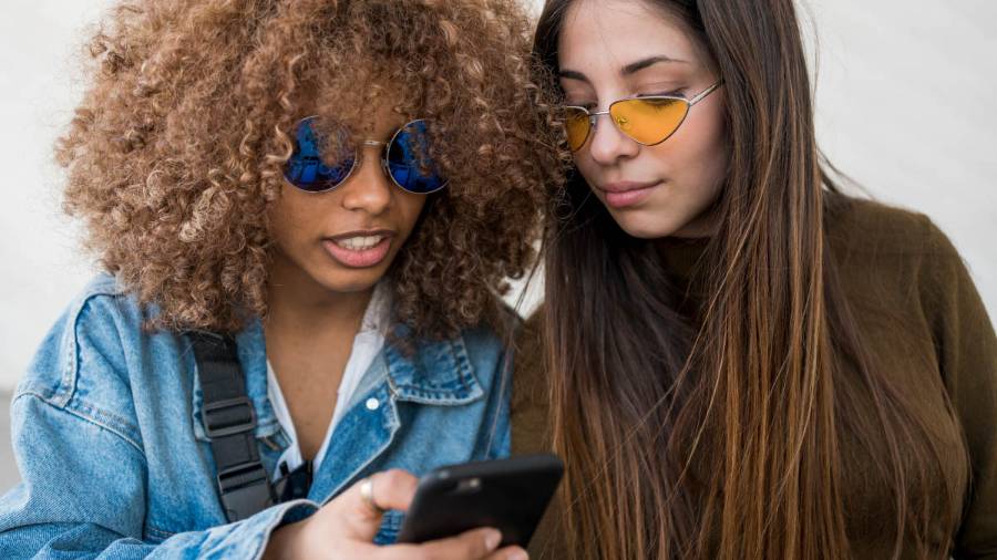 Two young people stand close together. One wears a denim jacket and tinted blue sunglasses, holding a phone. The other is in a brown top and tinted yellow-orange sunglasses, Both look at whatever is on the phone