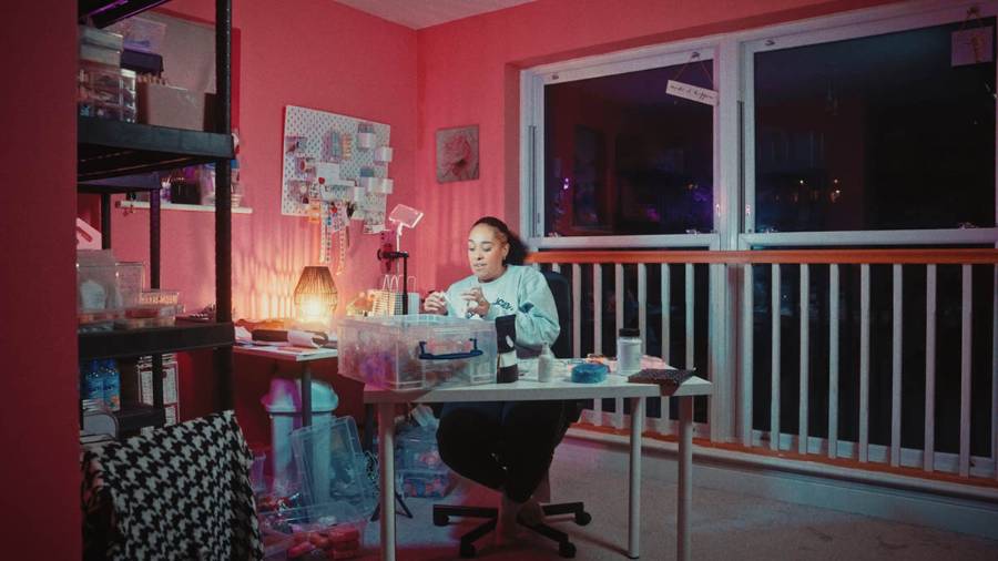 A person working at a desk in a pink-walled room, surrounded by tools, storage boxes, shelves and a desk lamp