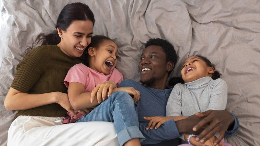 Two adults and two children laugh and play on a bed with light sheets, dressed in casual clothes