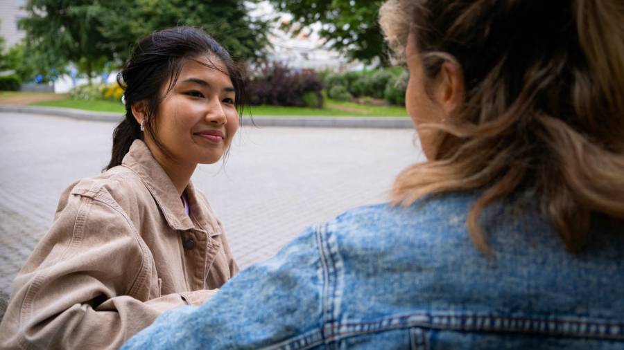 Two young people stand facing each other outside, one in a brown jacket and the other in denim, with trees and pavement in the background