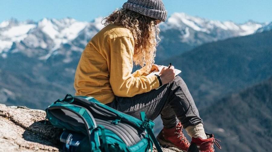 Woman dressed in mountain clothes, sat on mountain top taking notes with backpack beside her