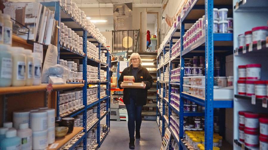 A person walking down an aisle in a store or warehouse, carrying several items, with shelves stocked with containers and bottles on both sides