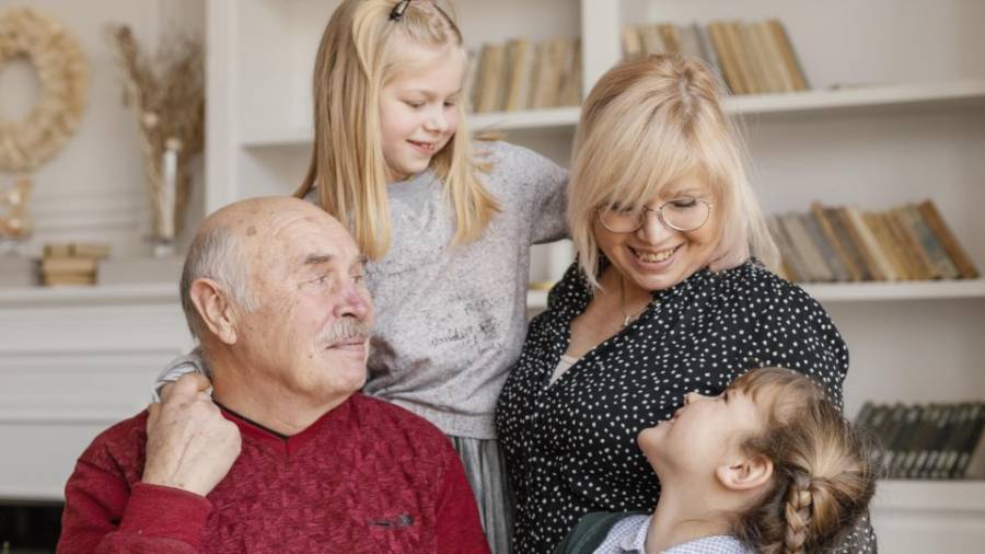 Four people standing closely together in front of a bookshelf filled with books and decorative items, including two adults and two children, who smile at each other