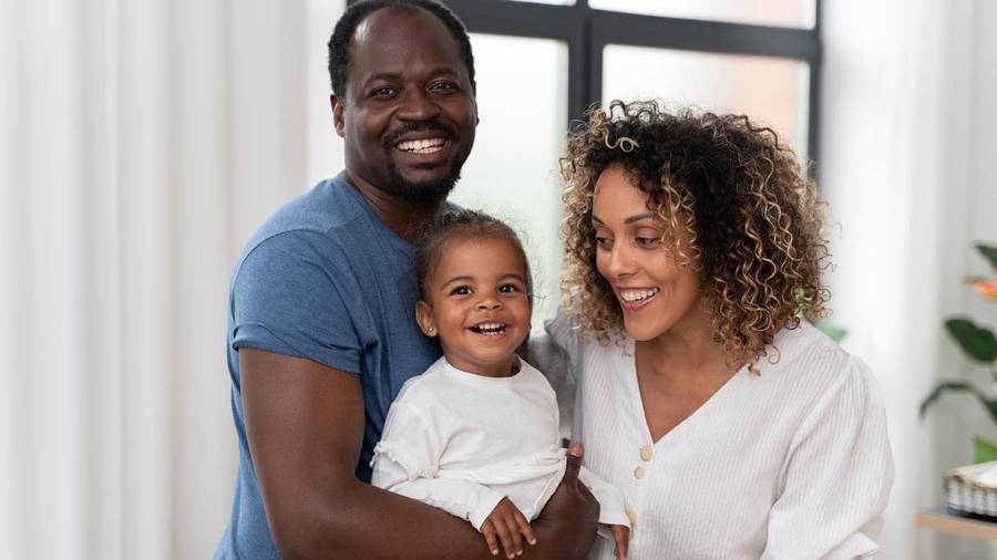 A couple stands indoors, with one adult holding a child while the other stands beside them, all dressed casually