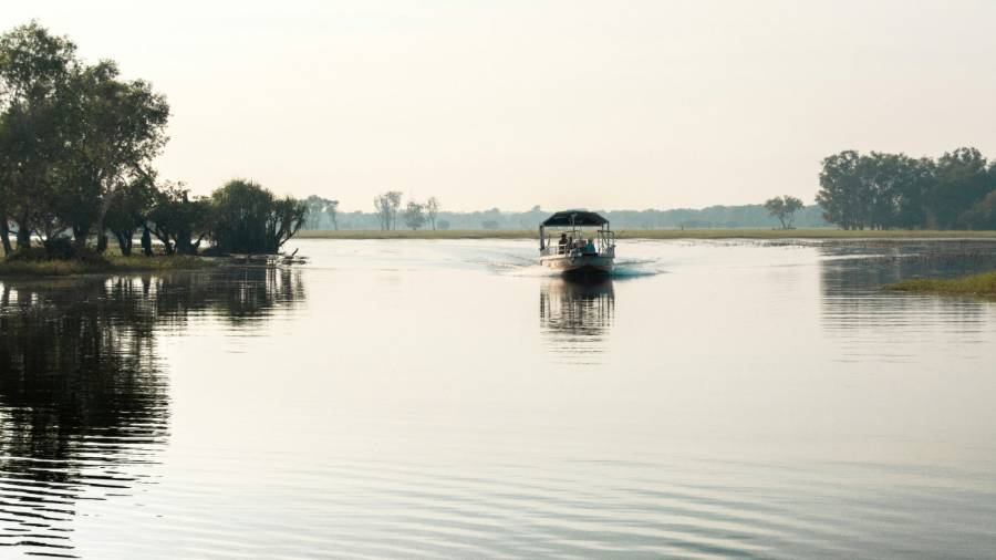 A small, covered boat navigates still, mirror-like waters in the Kakadu National Park in Australia’s Northern Territory. Hazy green riverbanks and trees are seen in the distance. The water reflects the banks and trees of nearby islands.