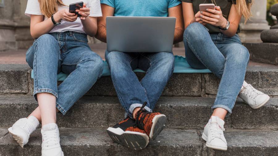 Three people in casual clothes sit on stone steps, one using a laptop, the others on their phones