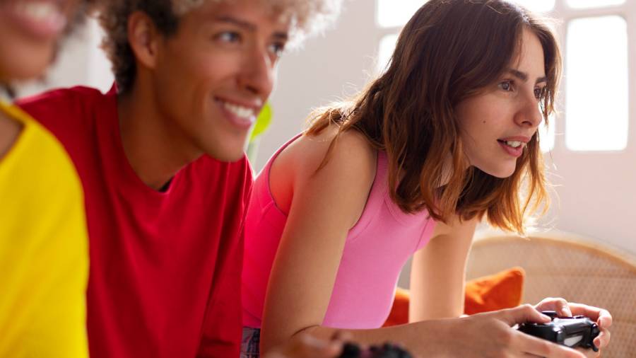 Three people sit close together, each holding a game controller. One wears yellow, one red and one a pink top