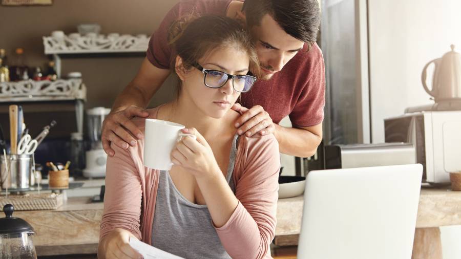 Two people at a kitchen table with a laptop, mug and paper documents, and shelves and utensils in the background