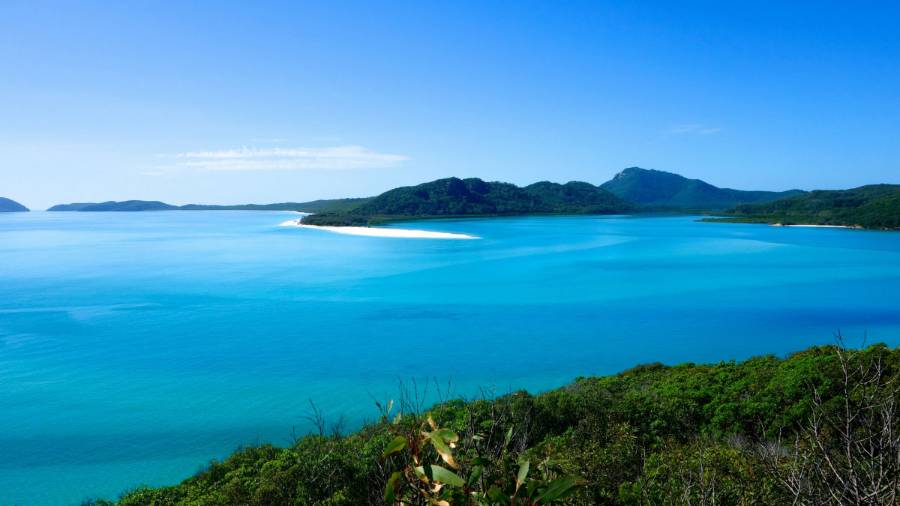 the ocean from the top of a green hill. The water is light blue, with a patch of pale white sand reaching into it from a land mass in the distance