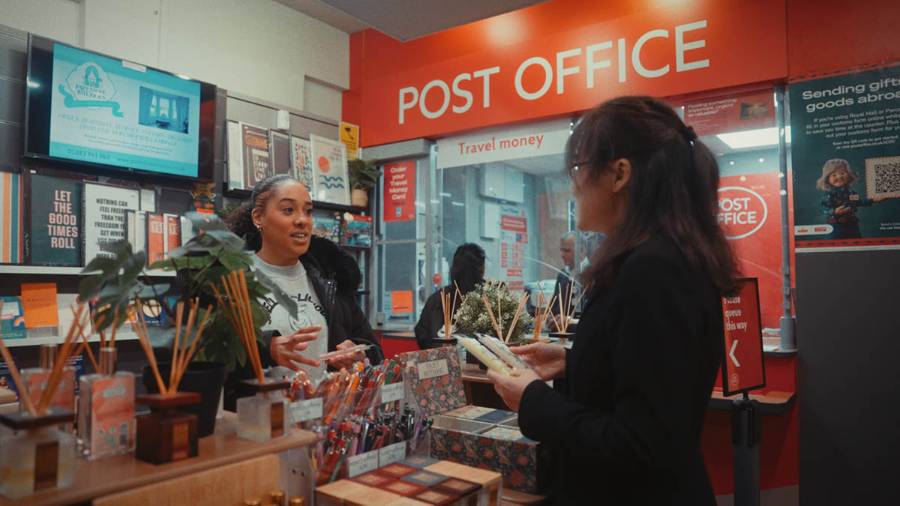 Two people interacting at an open Post Office counter, with stationery on shelves and a travel money counter behind