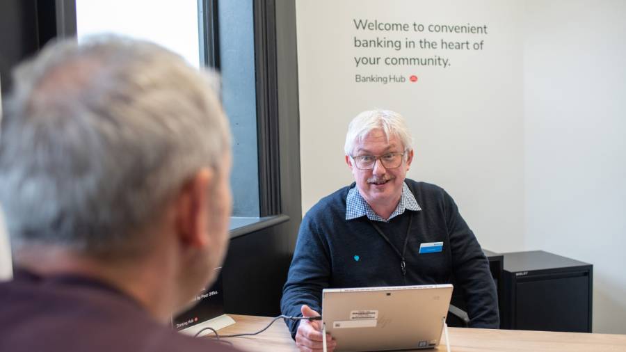 Two people sit across from each other at a desk in a bank-style setting. One wears a shirt, branded jumper and name tag. A wall sign behind them reads: 'Welcome to convenient banking in the heart of your community”