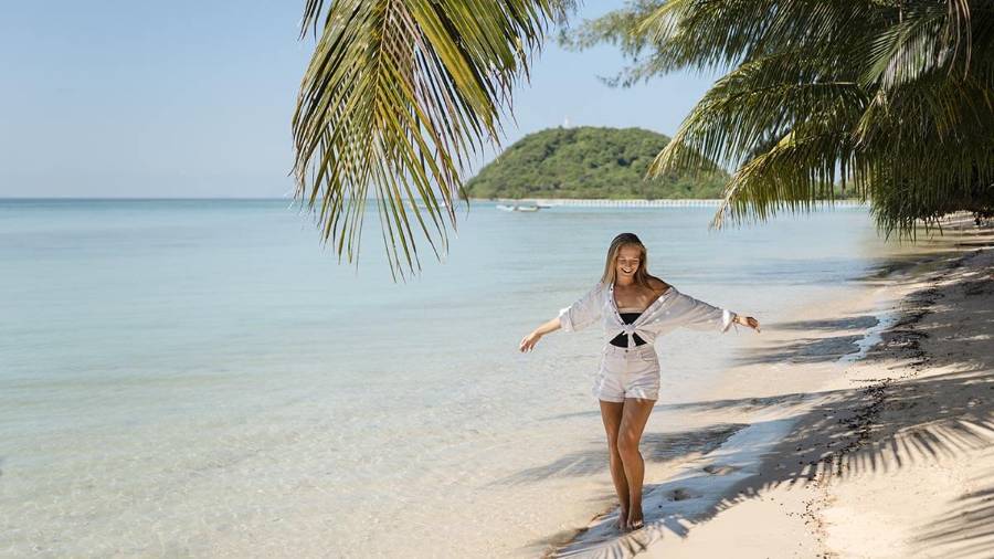 A person stands on a beach with palm trees, calm water and a distant island 
