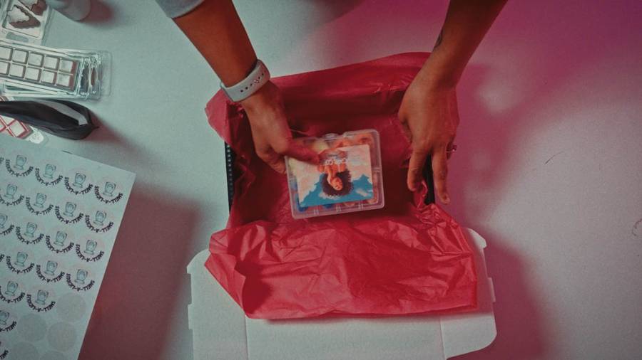 A person packing a small plastic container with an image of a woman into a box lined with red tissue paper, with stickers and packaging materials nearby