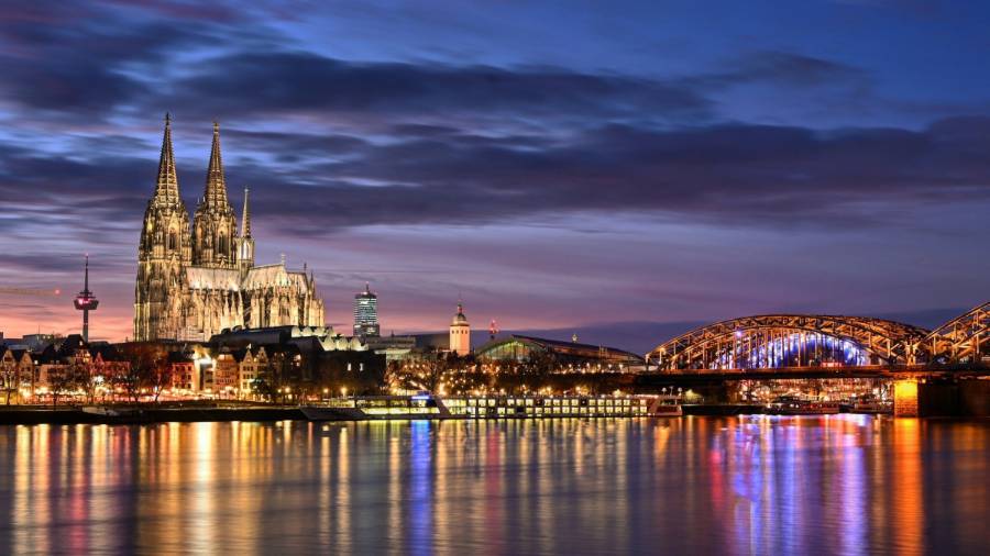 Lighted city buildings near body of water, including a cathedral with twin spires and a bridge spanning the water