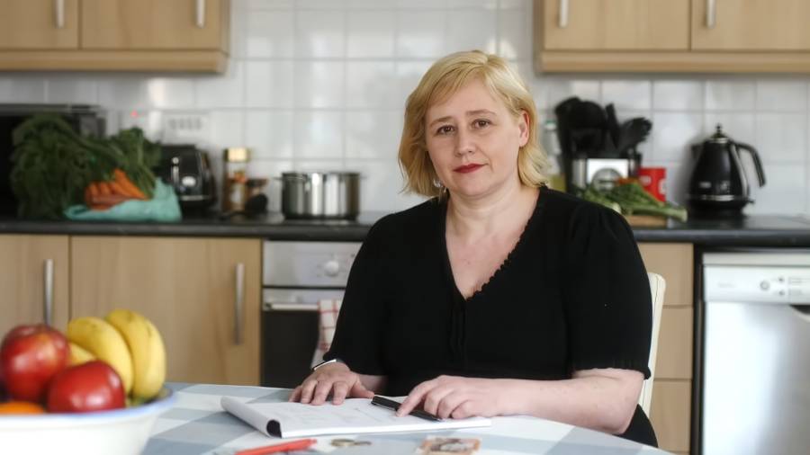 A blonde haired woman sits at the table in her kitchen, looking to camera and smiling contentedly