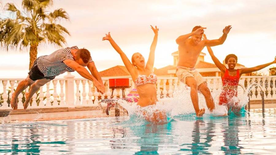 Two young men and two young women jumping in a pool with trees, houses and beach in distance