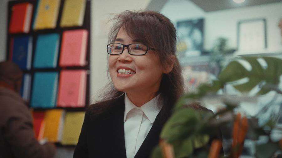 A smiling person in a black suit and white shirt indoors, with shelves of colourful books and plants in the background