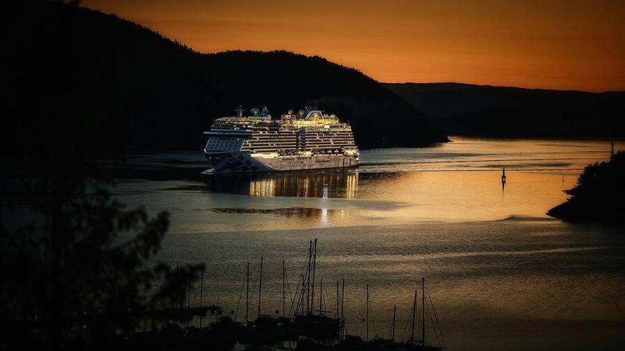 A lit cruise ship sails through a wider channel of water past hills at sunset 