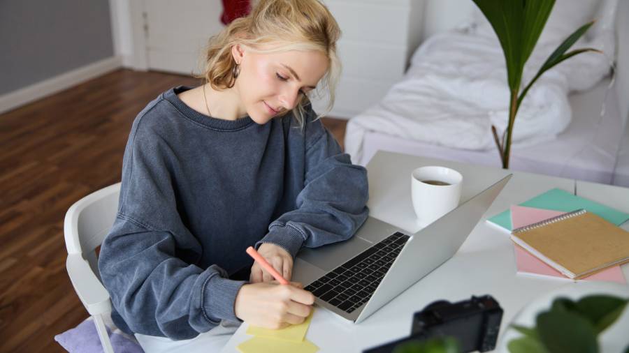 A person sits at a desk with a laptop, writing in a notepad, surrounded by coffee, notebooks and a camera, with a bed and plants in the background