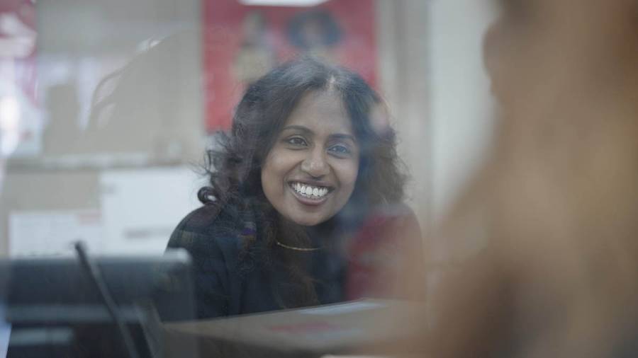 A person with curly hair behind a glass partition, with a computer and a red poster in the background