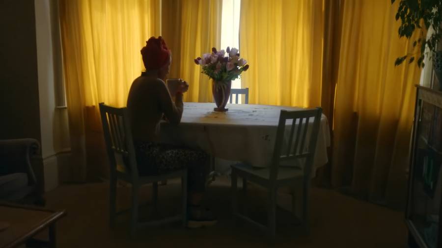 A woman sits at a dining table in a dark room with the curtains drawn, her back to the camera. She clutches a mug as though for comfort