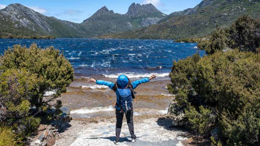 A hiker enjoying the natural beauty of Mount Cradle in the background and Dove Lake, an expanse of water marked with choppy waves, in Tasmania