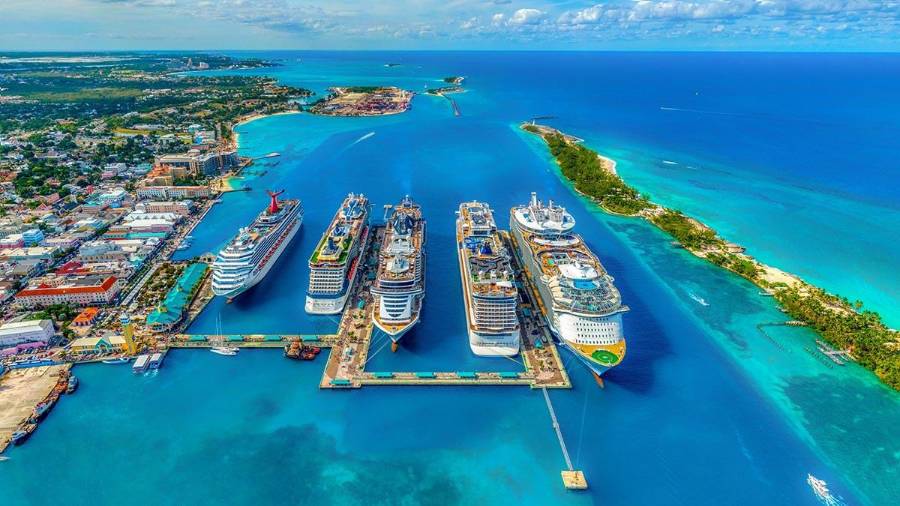 Aerial view of a port with six large cruise ships docked, clear blue water surrounding the area, and a strip of green land extending into the sea