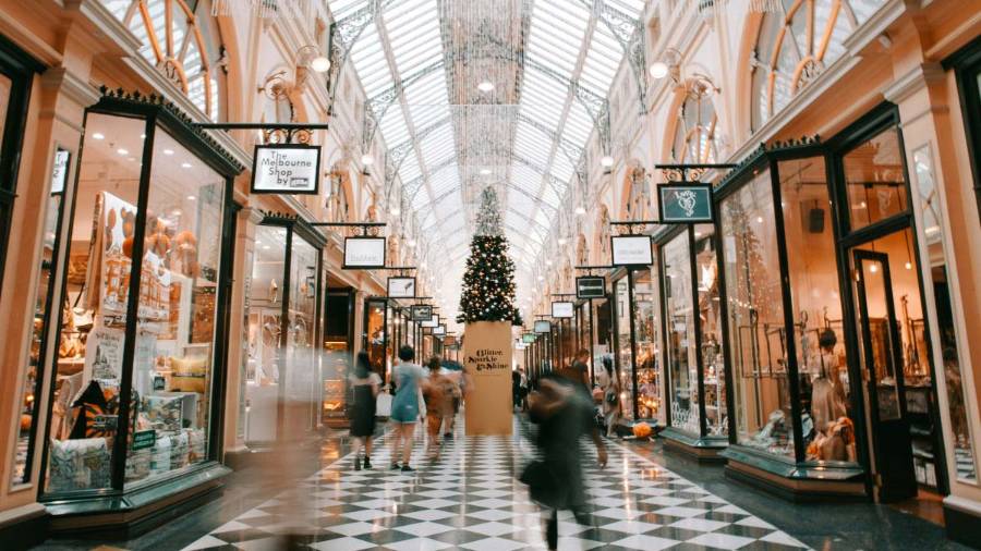 Shoppers walk through a shopping arcade. Above them is a pitched glass ceiling. Below their feet, a tiled floor is patterned with black and white diamonds. A tall Christmas tree reaches stands on a plinth in the background