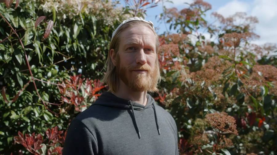 A bearded man in his thirties stands outdoors on a sunny day, his long hair pushed back by a hairband. He looks warmly towards the camera