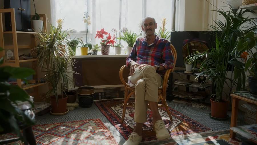 Someone’s in a room full of clothes aA man sits in a wooden chair in his home, surrounded by houseplants and smiling to cameraand accessories, using a tablet while surrounded by shelves and racks of fashion gear