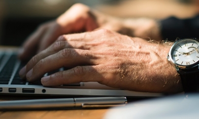 A man's hand, wearing a watch, typing on his laptop on his desk