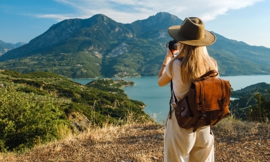 Woman taking picture of landscape featuring a mountain range and lake.