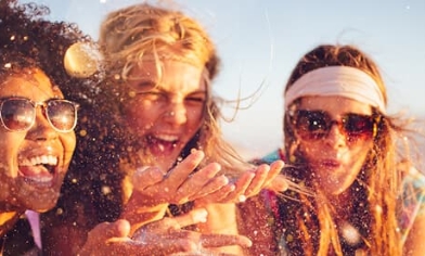 Three young women, left and right both wearing sunglasses, all laughing in the sunshine