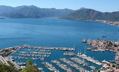 a marina with rows of boats and yachts docked along a calm coastline, surrounded by mountainous terrain and nearby buildings