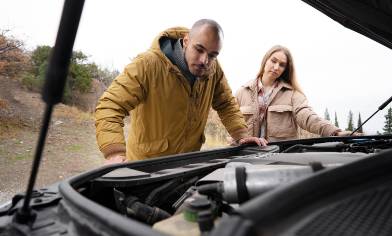 A couple stands by the open bonnet of a presumably broken-down car, looking down at the engine inside