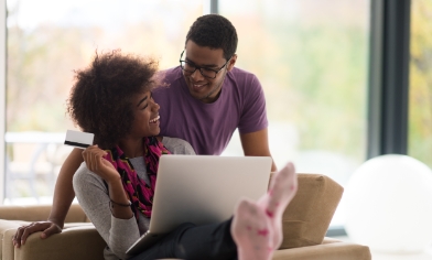 Woman and partner laughing together, whilst she is sitting down with laptop, holding a credit card in one hand