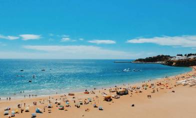 Raised view of a sandy beach where a light sea and sky. The beach is dotted with umbrellas, blankets and people enjoying their holidays. In the distance, small white buildings and dark vegetation line the shore. 