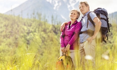 Older couple with backpacks looking to distance on green hillside with mountains in background