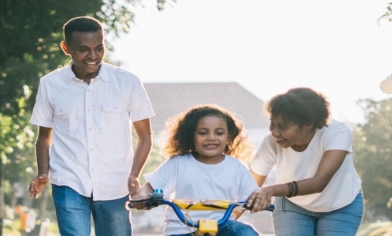 Mother and father teaching their little girl to ride a bike along a sunlit road