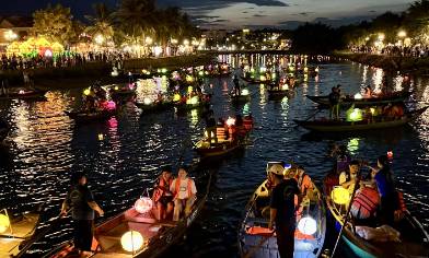 Boats with lanterns float on a river at night, with crowds watching from lit-up riverbanks.